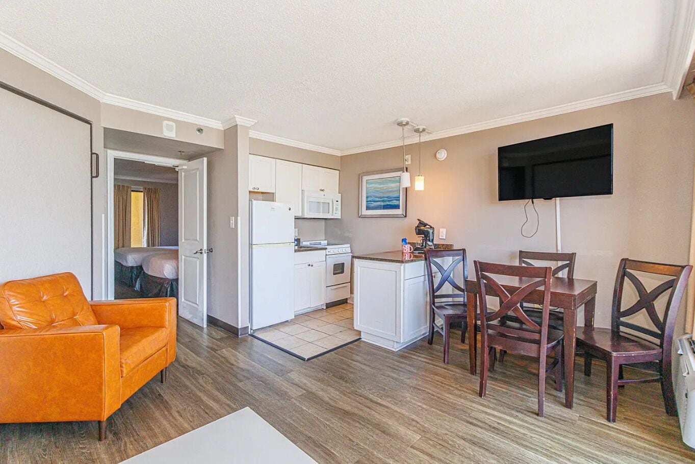 A compact living space with a kitchenette, a dining area with wooden chairs, a sofa, and a flat-screen TV mounted on the wall.