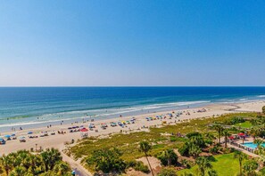 Ein Strand mit Sonnenschirmen, Menschen, einem Schwimmbad und Palmen.