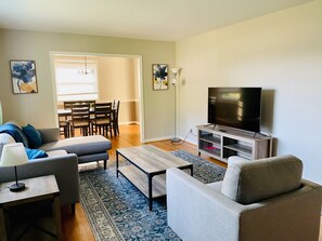 A living room with a sofa, a coffee table, a TV on a stand, and a dining area with chairs and a table. - Peaceful 4-bedroom ranch in Richmond’s west end. (Richmond)