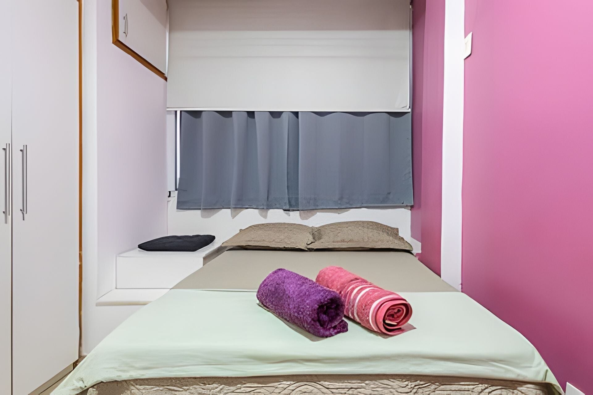 Una piccola stanza con un letto singolo e un letto a castello, una finestra con tende grigie, e un letto con copriletto verde e due asciugamani arrotolati.