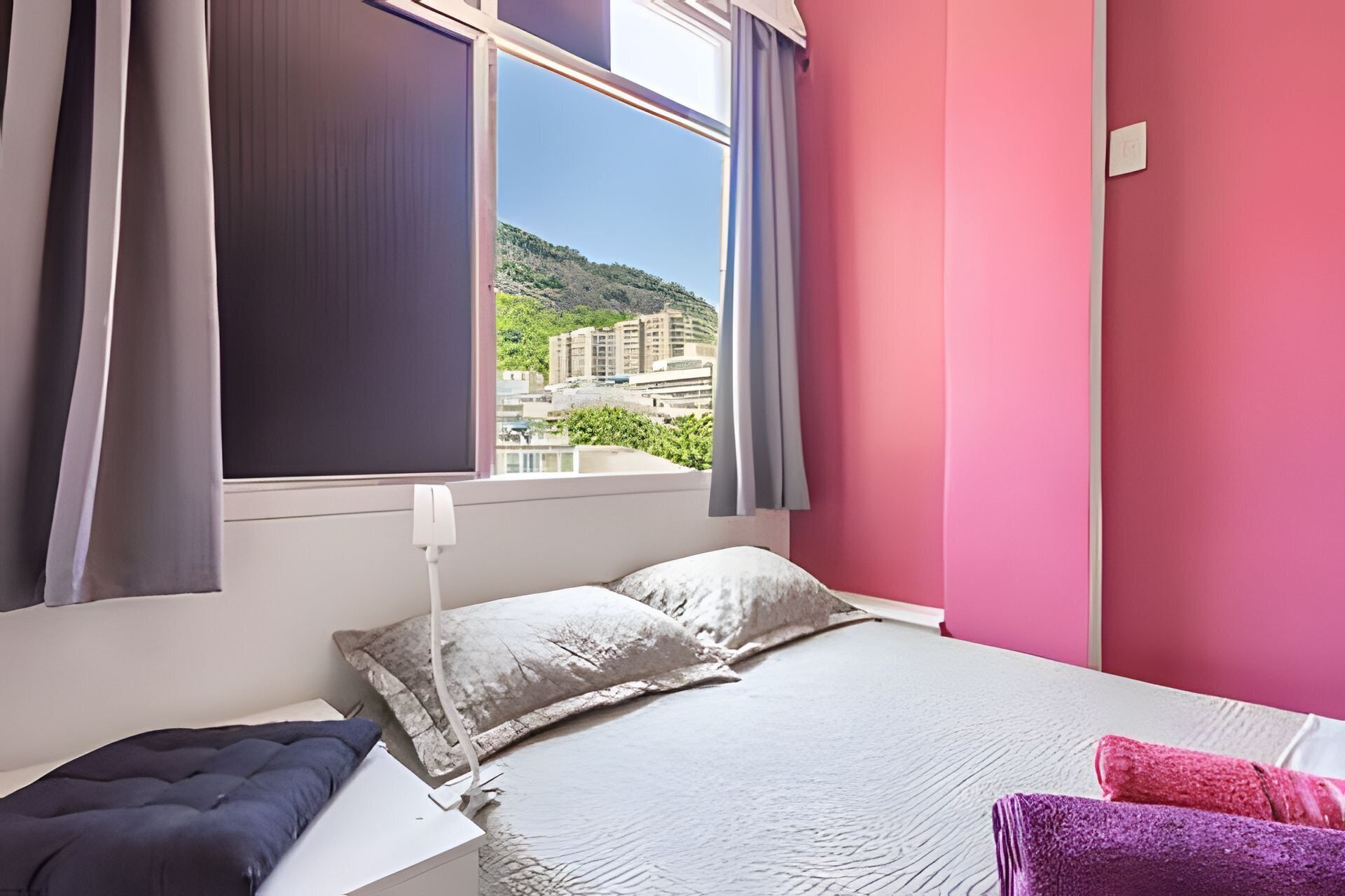 A bedroom with a bed, pillows, a bedside table, a window with curtains, and a view of buildings and greenery.