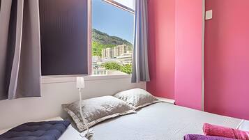 Quarto com cama, almofadas, mesa de cabeceira, janela com cortinas e vista para edifícios e áreas verdes.