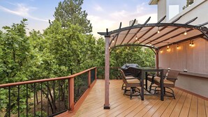 Une terrasse en bois agrémentée d’une pergola, d’un espace salon extérieur et offrant une vue sur une zone boisée.