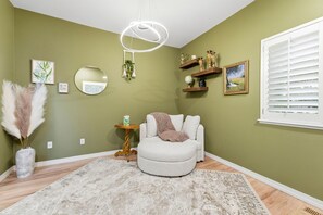 Ein Zimmer mit einem runden Spiegel, einem runden Hocker, einem kleinen Tisch mit einer Vase und einem Fenster mit Jalousien.