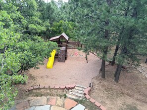 Ein Spielplatz mit Rutsche und Kletterbereich, umgeben von Bäumen und einem hölzernen Spielhäuschen.