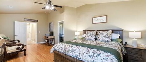 A bedroom with a bed, bedside tables, a desk, a chair, a ceiling fan, and a door.