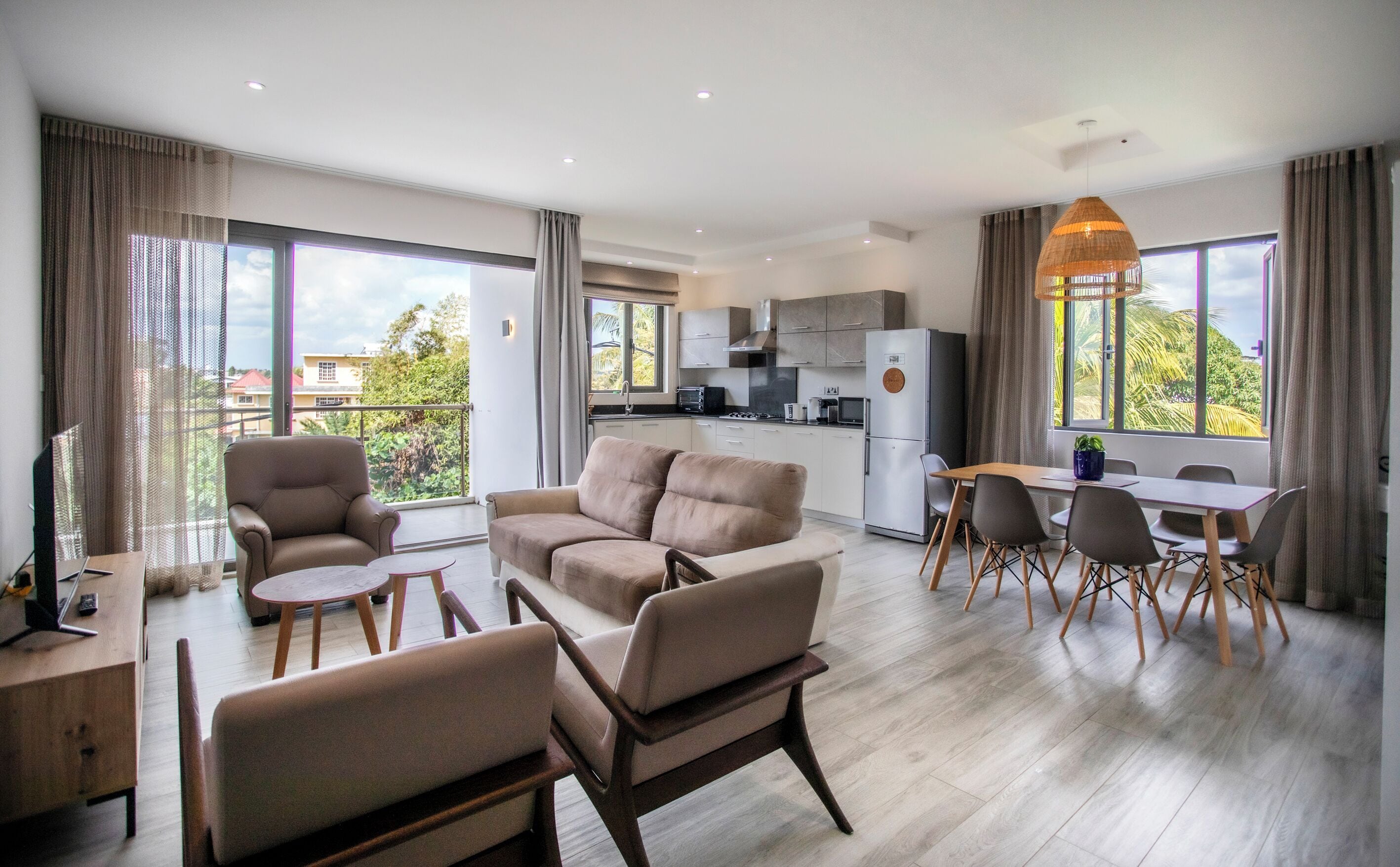 A modern living room with a sofa set, a dining area with a table and chairs, a kitchen with appliances, and large windows with curtains.