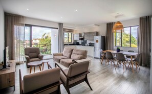 Un salon moderne comprenant un ensemble de canapés, un coin repas avec une table et des chaises, une cuisine équipée et de grandes fenêtres avec des rideaux.