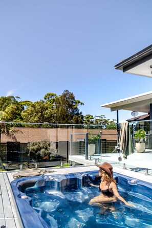 A person in a hot tub on a rooftop with a clear sky, surrounded by trees and a modern building. - Summer and Winter by The Sea - Outdoor Spa (Mollymook Beach)