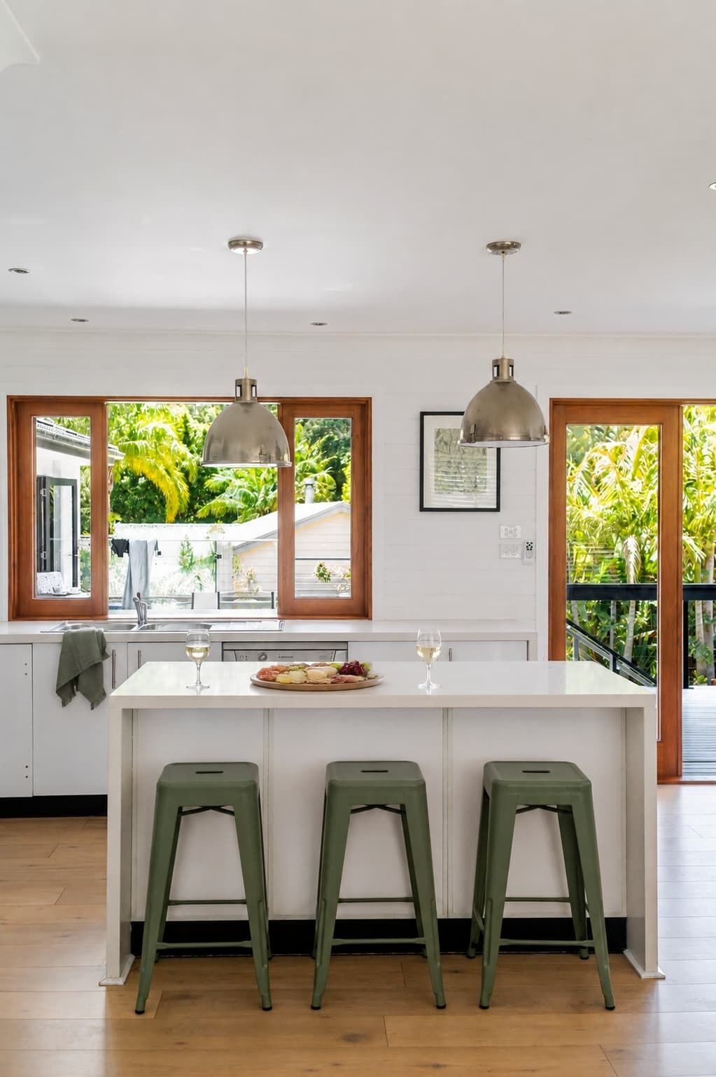 A modern kitchen with a central island, two hanging pendant lights, and a view of greenery outside.