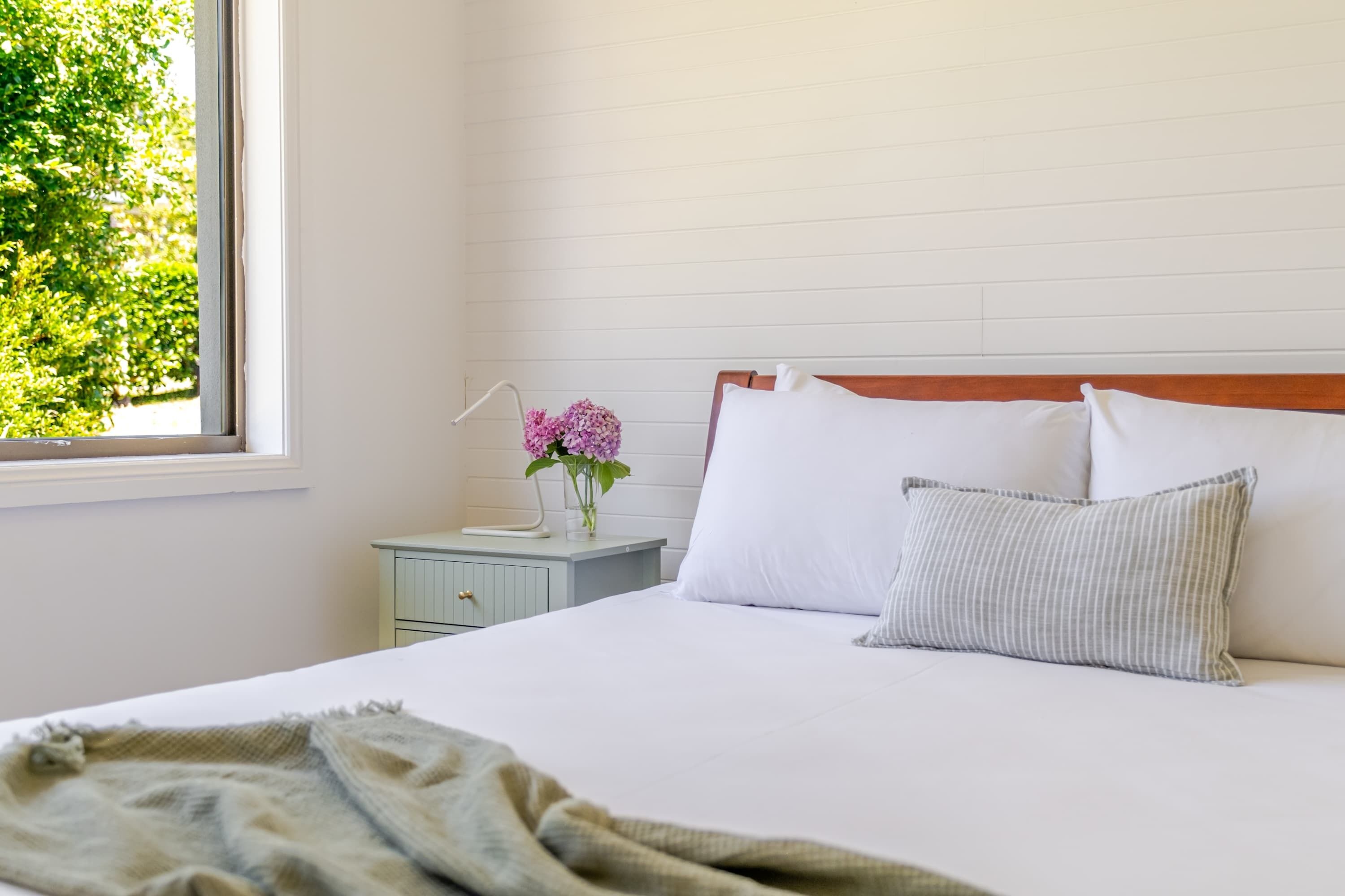 A bedroom with a bed, a nightstand, a window, and a view of greenery outside.
