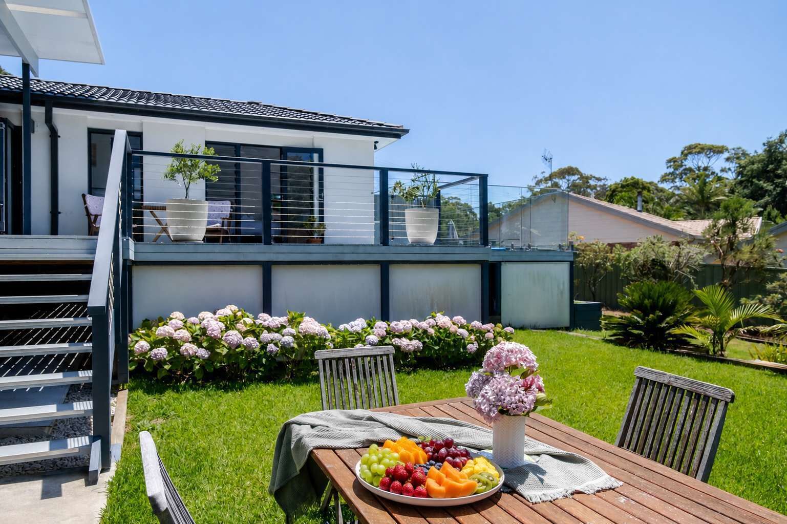 A modern house with a balcony, a wooden table with a fruit platter, and a garden with hydrangeas.