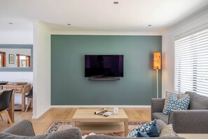 Una sala moderna con un televisor de pantalla plana montado en una pared verde azulada, una mesa de centro de madera, un sofá gris con cojines estampados y una ventana con persianas blancas.