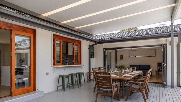 Un área de comedor al aire libre con una mesa preparada para cuatro, rodeada de sillas y taburetes de madera, con vista al interior a través de puertas de vidrio.