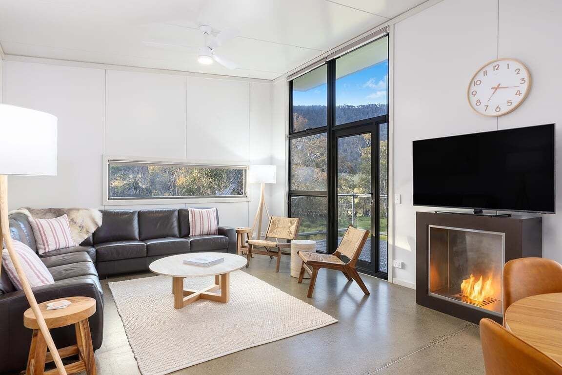 A modern living room with a fireplace, a sofa, chairs, a TV, and a large window offering a view of the outdoors.