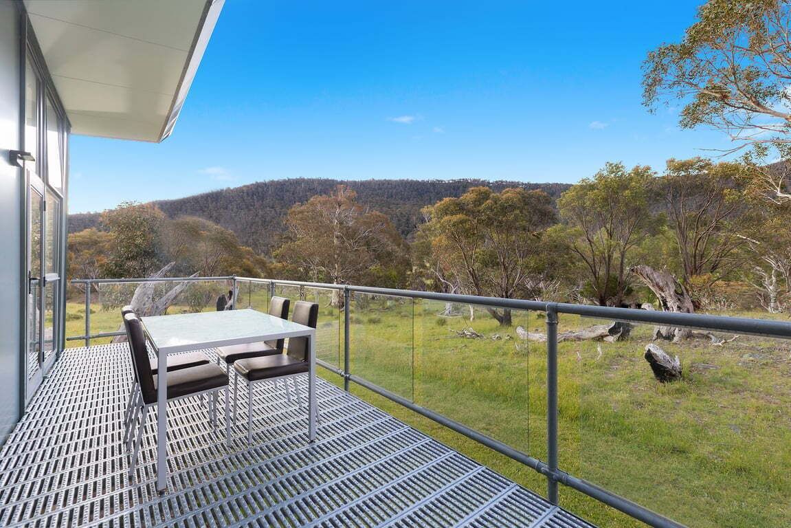 Een balkon met een metalen rooster vloer, een tafel en stoelen, met uitzicht op een grasachtig landschap met bomen en heuvels.