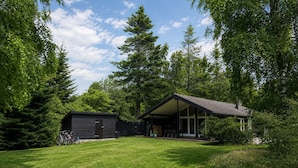 Ein modernes Haus mit überdachter Terrasse und kleinem Gartenhaus, umgeben von dichten Bäumen.