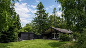Ein modernes Haus mit überdachter Terrasse und kleinem Gartenhaus, umgeben von dichten Bäumen.