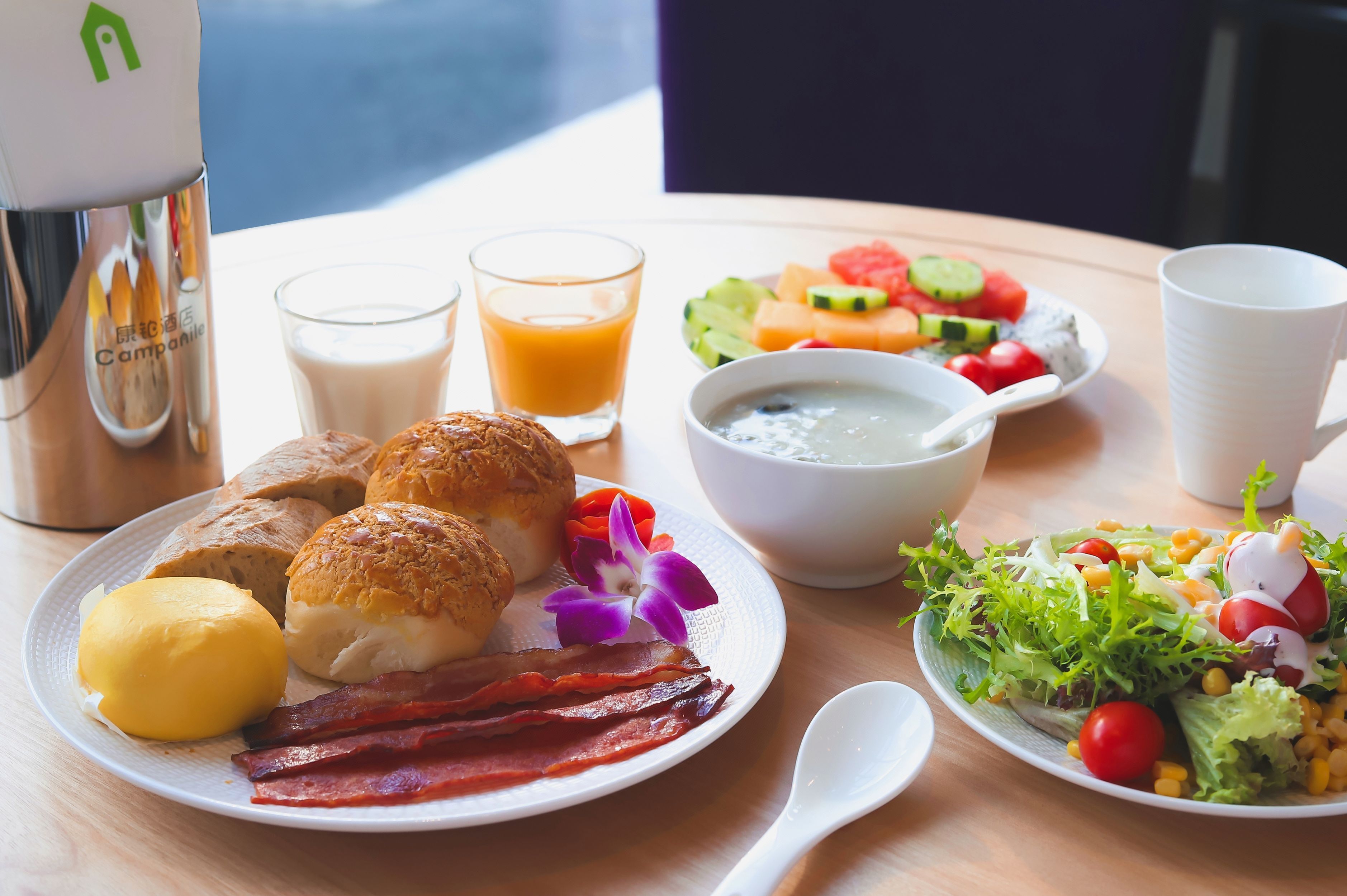 A breakfast setting with bread, eggs, bacon, a bowl of soup, a fruit salad, and a cup of coffee.