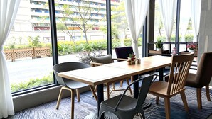 Uma sala de jantar moderna com mesa e cadeiras, janelas amplas com cortinas e vista para um edifício com varandas.