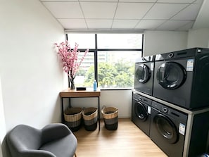 A laundry room with two stacked washing machines, a chair, a small table with a vase of flowers, and baskets.