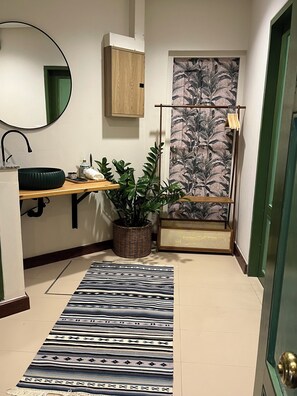 A bathroom with a wooden cabinet, a mirror, a sink, a potted plant, a patterned rug, and a door with a green frame. - Welcome to Chur’s Garden Boutique Stay 100-Year-Old Heritage Chinatown Bangkok (Krung Thep Maha Nakhon)