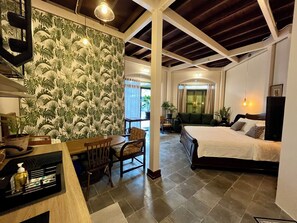 Quarto de hotel com cama, área de estar com mesa e cadeiras, janela grande com cortinas e parede com padrão de folhagem tropical.
