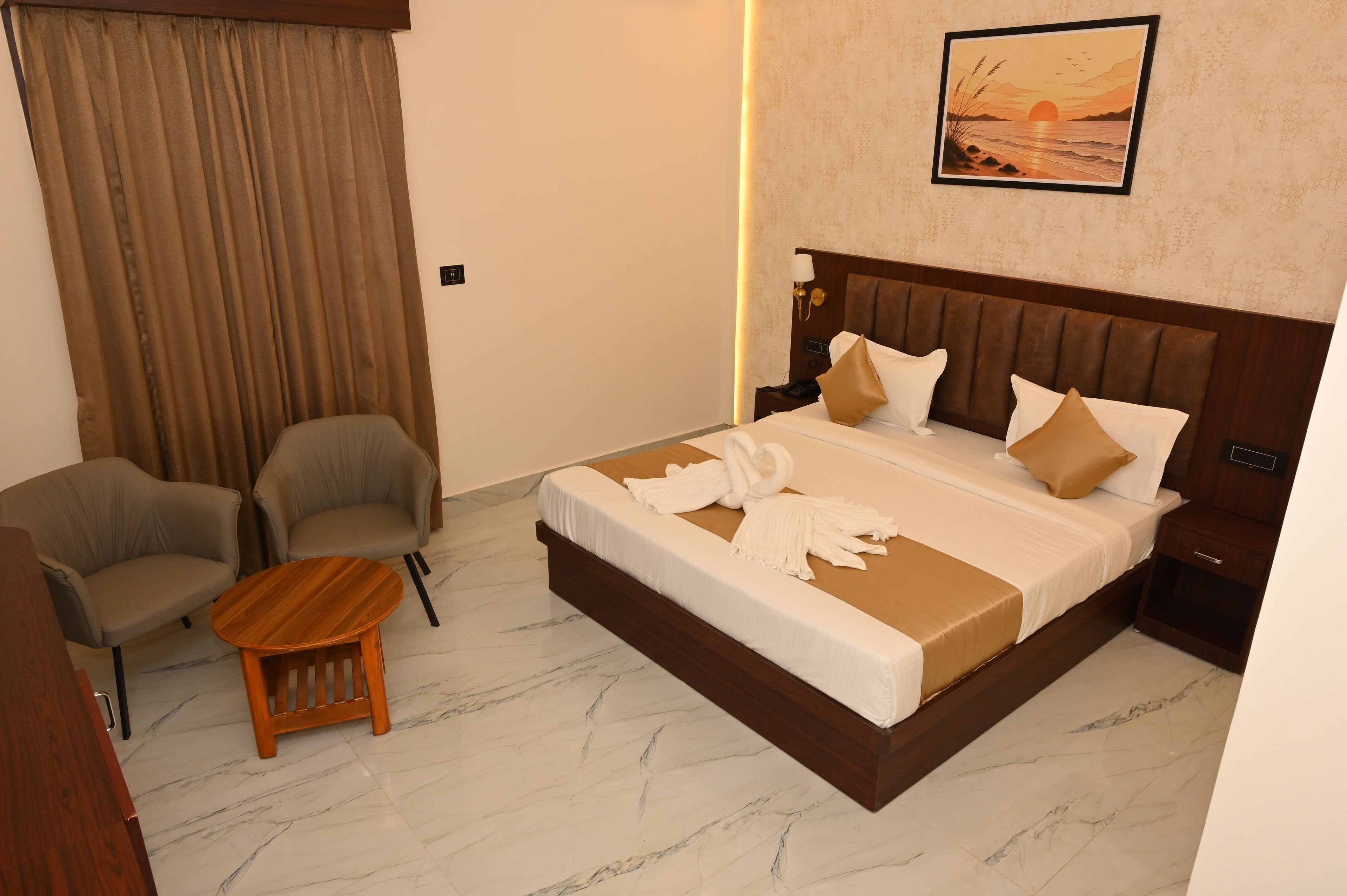 A hotel room with a large bed, two armchairs, a small wooden table, and a painting of a sunset on the wall.