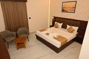 A hotel room with a large bed, two armchairs, a small wooden table, and a painting of a sunset on the wall. - BENGALURU VISTA  (Ayodhya)