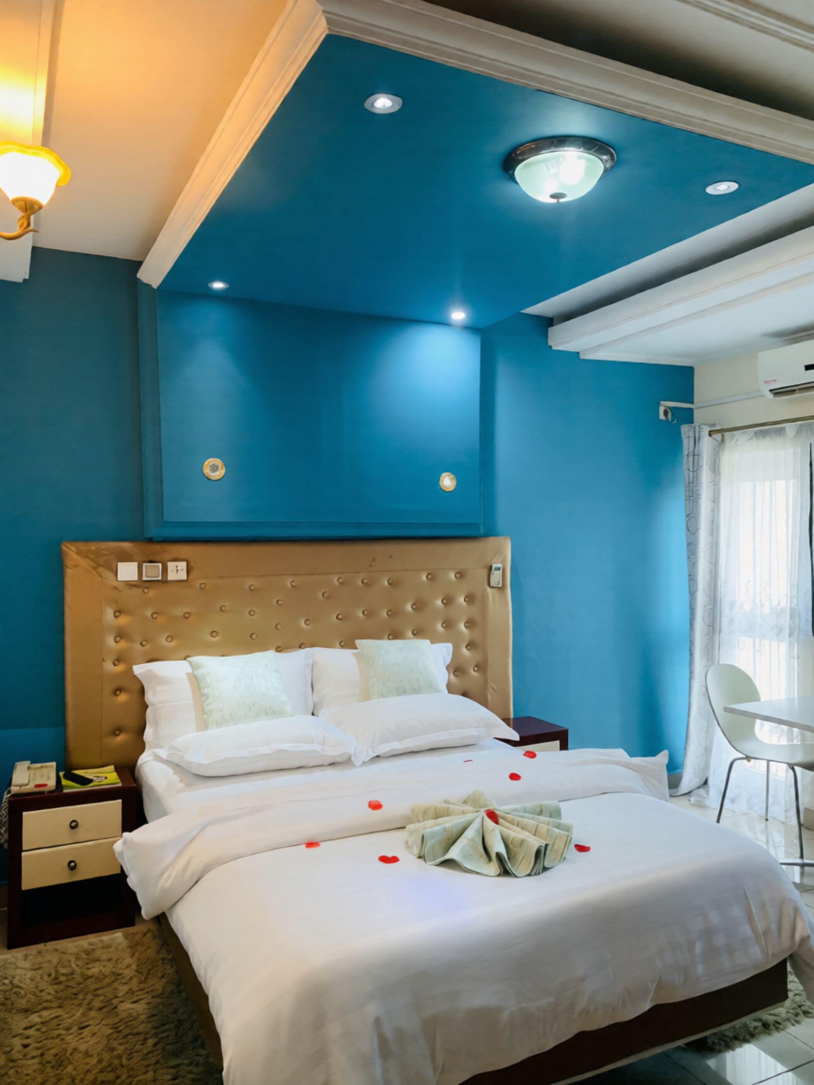 A hotel room with a large bed, a headboard, a bedside table, a nightstand, a lamp, a window with curtains, and a ceiling light.