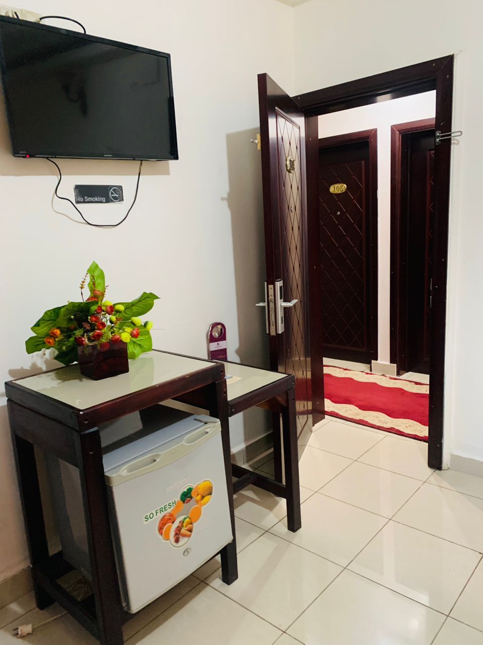 A room with a television mounted on the wall, a table with a plant, a refrigerator, and a door with a number plate.
