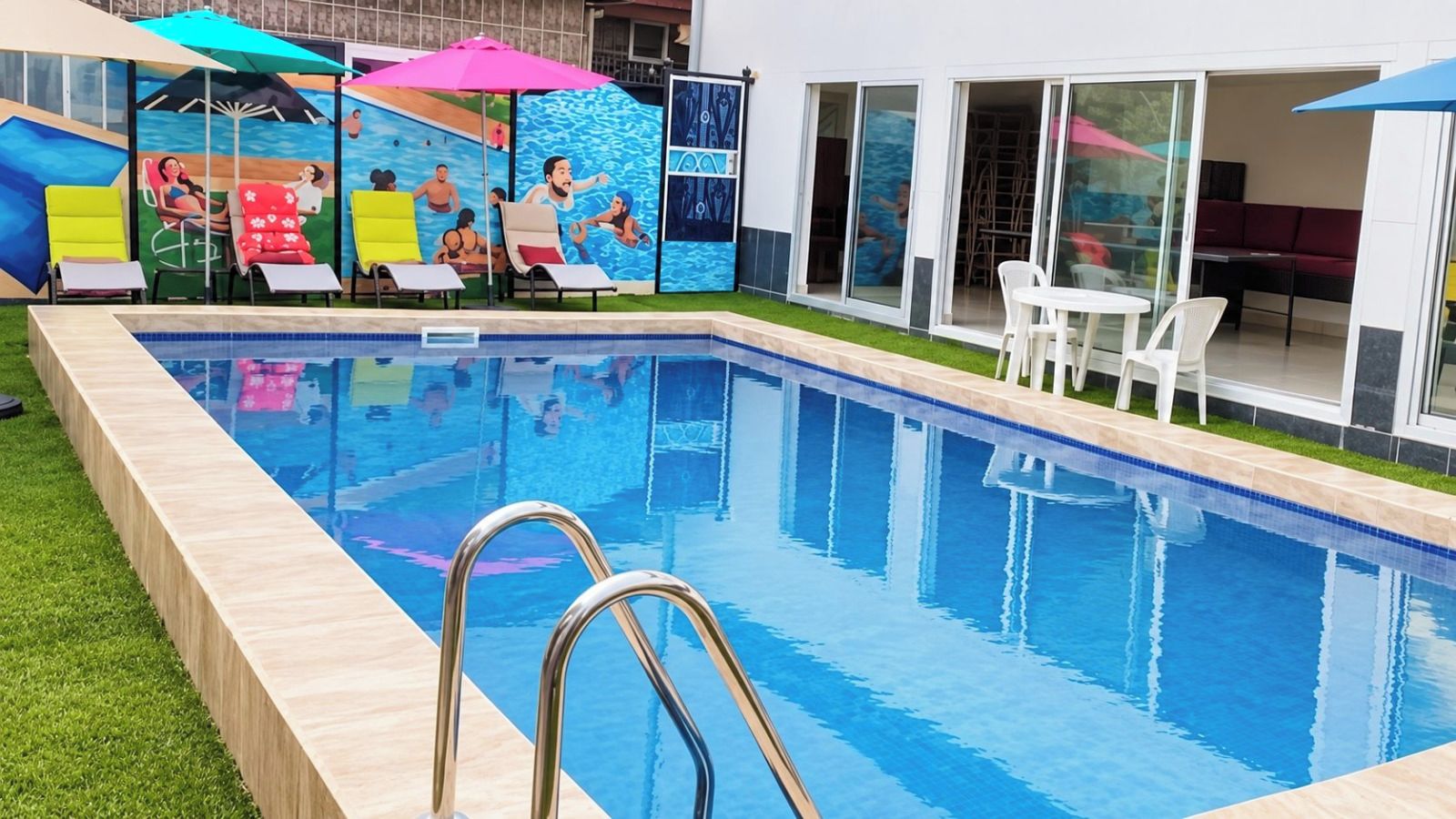 Una piscina con superficie de agua azul cristalina, rodeada de césped y un edificio con puertas de vidrio.