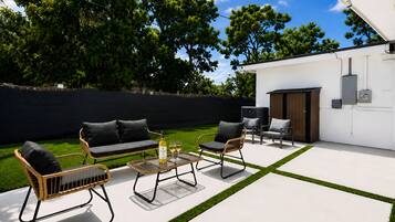 A modern outdoor seating area with a sofa, chairs, and a table, set against a white wall and surrounded by greenery.