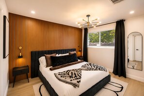 Un dormitorio moderno con pared de paneles de madera, cabecera tapizada de color negro, cama grande con ropa de cama estampada, lámparas de noche y una lámpara de techo.