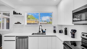 A modern kitchen with white cabinetry, a stainless steel dishwasher, a built-in oven, and a window with blinds.