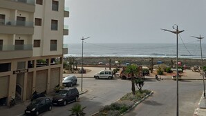 Un edificio a più piani con un'area di parcheggio, una strada con auto parcheggiate e vista sul mare.