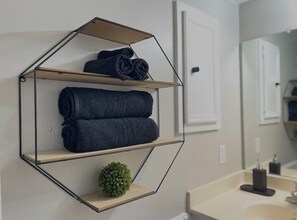 A bathroom with a hexagonal towel rack holding rolled towels, a small potted plant, and a mirror reflecting the room. - Golf Suites by Reserved Rentals (Lubbock)