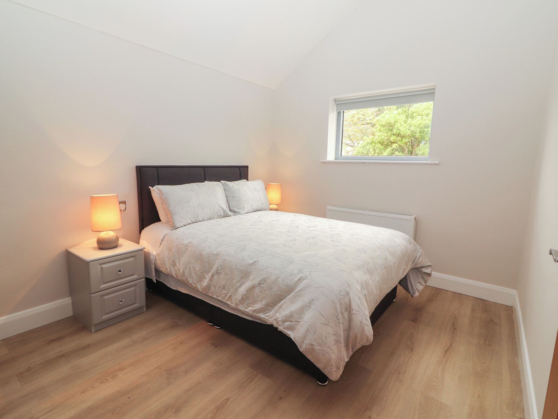 Un dormitorio con cama, mesitas de noche, ventana y radiador.