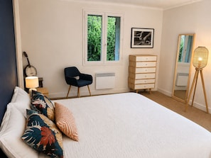 A bedroom with a bed, a chair, a dresser, a lamp, a guitar, and a window with a view of greenery. - House in Yvoire between Lake and Village (Yvoire)