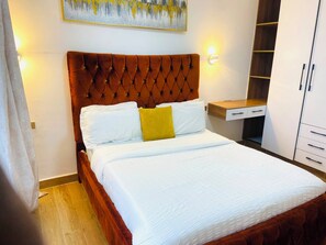 A hotel room with a bed, a wooden headboard, a nightstand, a wardrobe, and a painting on the wall. - Cozy 1-bedroom apartment in lovely Kilimani with gym (Nairobi)