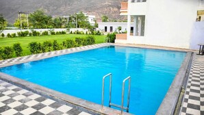 En swimmingpool med ett rutmönstrat kantparti, metallstegar och utsikt över en vit byggnad och grönskande växtlighet i bakgrunden.