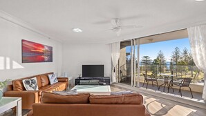 Ein modernes Wohnzimmer mit einer Couch, einem Couchtisch und einem Flachbildfernseher. Ein Balkon bietet Blick auf Bäume und einen wolkenlosen Himmel.