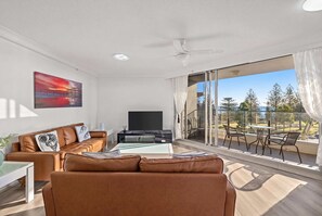 A modern living room with a sofa, a coffee table, and a flat-screen TV. There is a balcony with a view of trees and a clear sky. - "HAVEN" By TheGuestHouse. (Broadbeach)