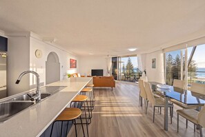 A modern kitchen with a central island, bar stools, a dining area with a glass table, and a view of the ocean. - "HAVEN" By TheGuestHouse. (Broadbeach)