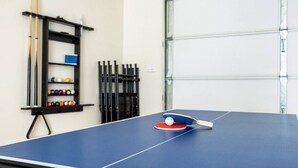 Een tafeltennistafel met batje en bal, een biljartbalhouder en een poolbiljart in een ruimte met een garagedeur.