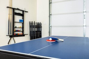 Ein Tischtennistisch mit Schläger und Ball, ein Billardballständer und ein Billardtisch in einem Raum mit einer Garagentür.