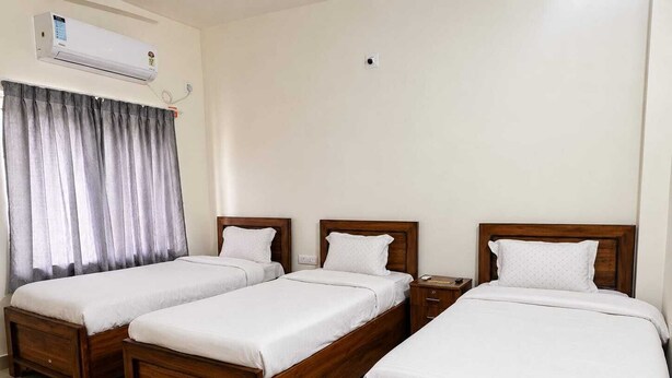 A hotel room with three wooden beds, a red and beige rug, a window with grey curtains, and an air conditioning unit on the wall. - A Boutique Hotel (Bhubaneshwar)