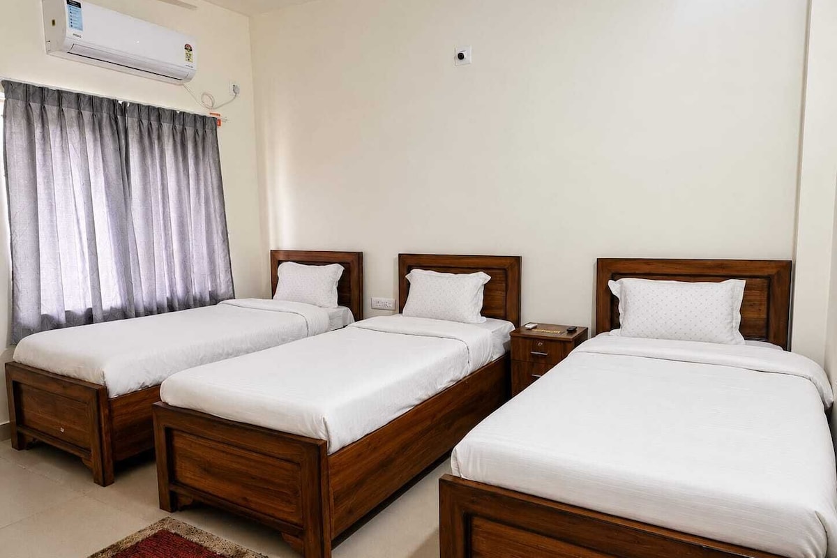 A hotel room with three wooden beds, a red and beige rug, a window with grey curtains, and an air conditioning unit on the wall.
