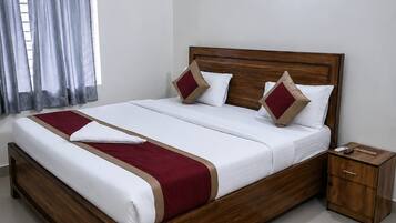 Una cama bien hecha con estructura de madera y ropa blanca, una mesita de noche con lámpara, y una alfombra estampada en el suelo.