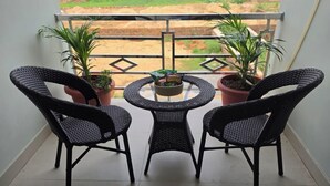 Un balcón con dos sillas de mimbre y una mesa redonda pequeña, rodeado de plantas en macetas.